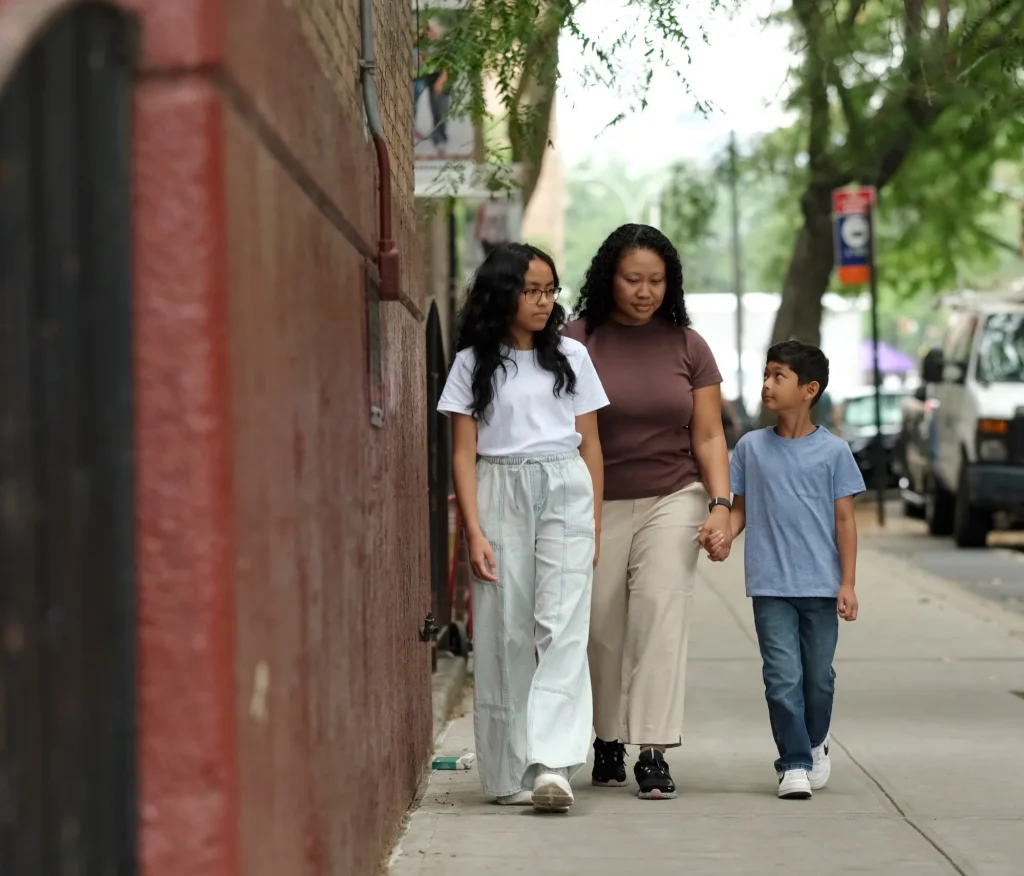 A mother walks along a sidewalk holding hands with her son and daughter