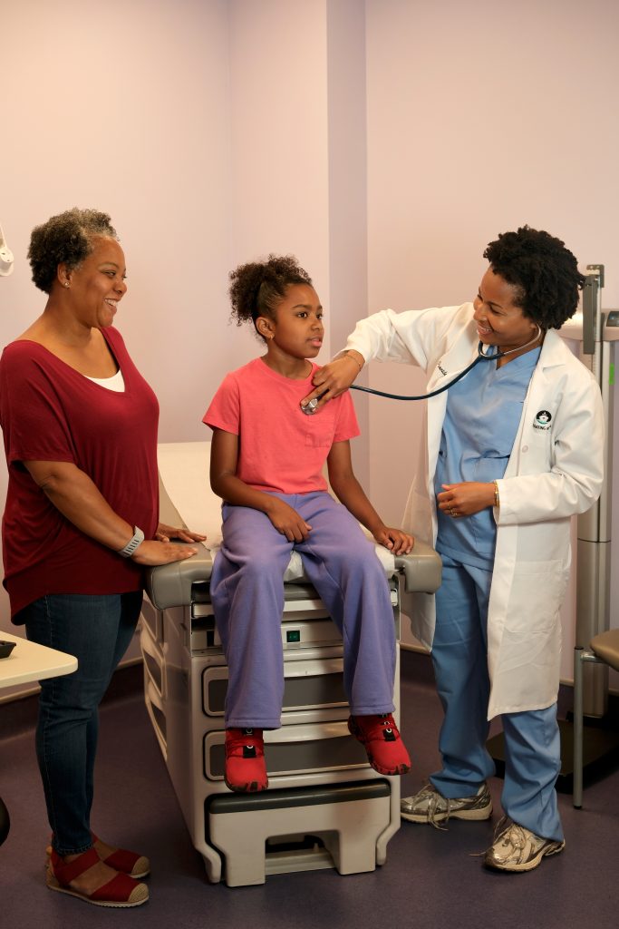 A doctor listens to a young patient's heartbeat with a stethoscope while her mother watches